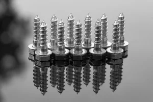 A Row Of Metal Carriage Bolts With Hexagonal Heads Placed Upright With Their Reflections Visible On A Polished Surface.